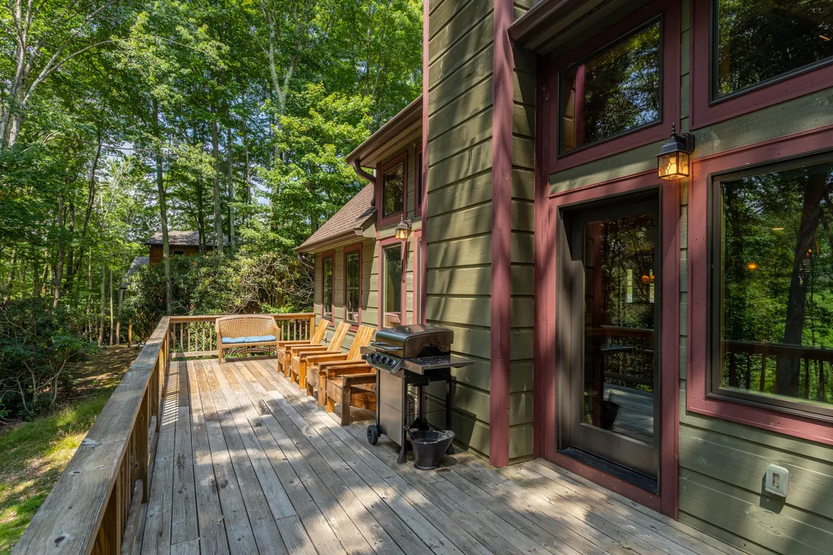 An inviting deck furnished with comfortable seating and a grill, surrounded by trees and featuring large windows that connect the interior to the outdoors. The wooden railing and leafy backdrop set a tranquil tone for relaxing or socializing.