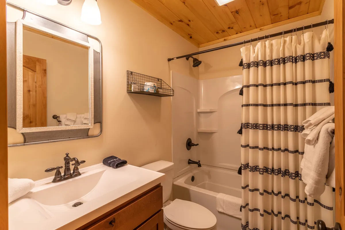 A well-appointed bathroom with a vanity, sink, mirror, and shower with curtain beneath a wooden ceiling. The space feels relaxing and fresh, providing the convenience described as part of the lower level amenities.