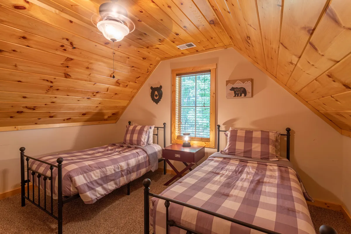 A charming twin bedroom with plaid bedding, a wooden ceiling, bear-themed wall decor, and a window letting in natural light. The space feels fun and welcoming, perfect for kids or friends to share.