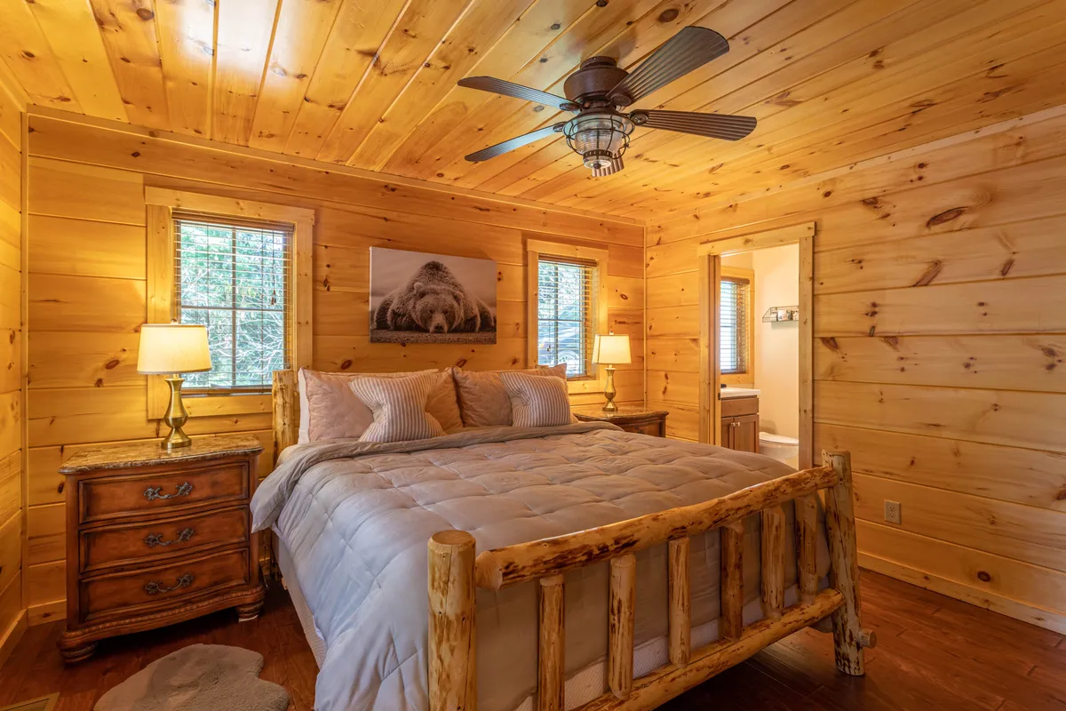 A tranquil bedroom featuring a rustic log bed flanked by wooden nightstands, with framed wildlife art on the wall and a window offering views of the surrounding nature. Soft, warm lighting creates a peaceful atmosphere ideal for relaxation or enjoying a quiet morning.