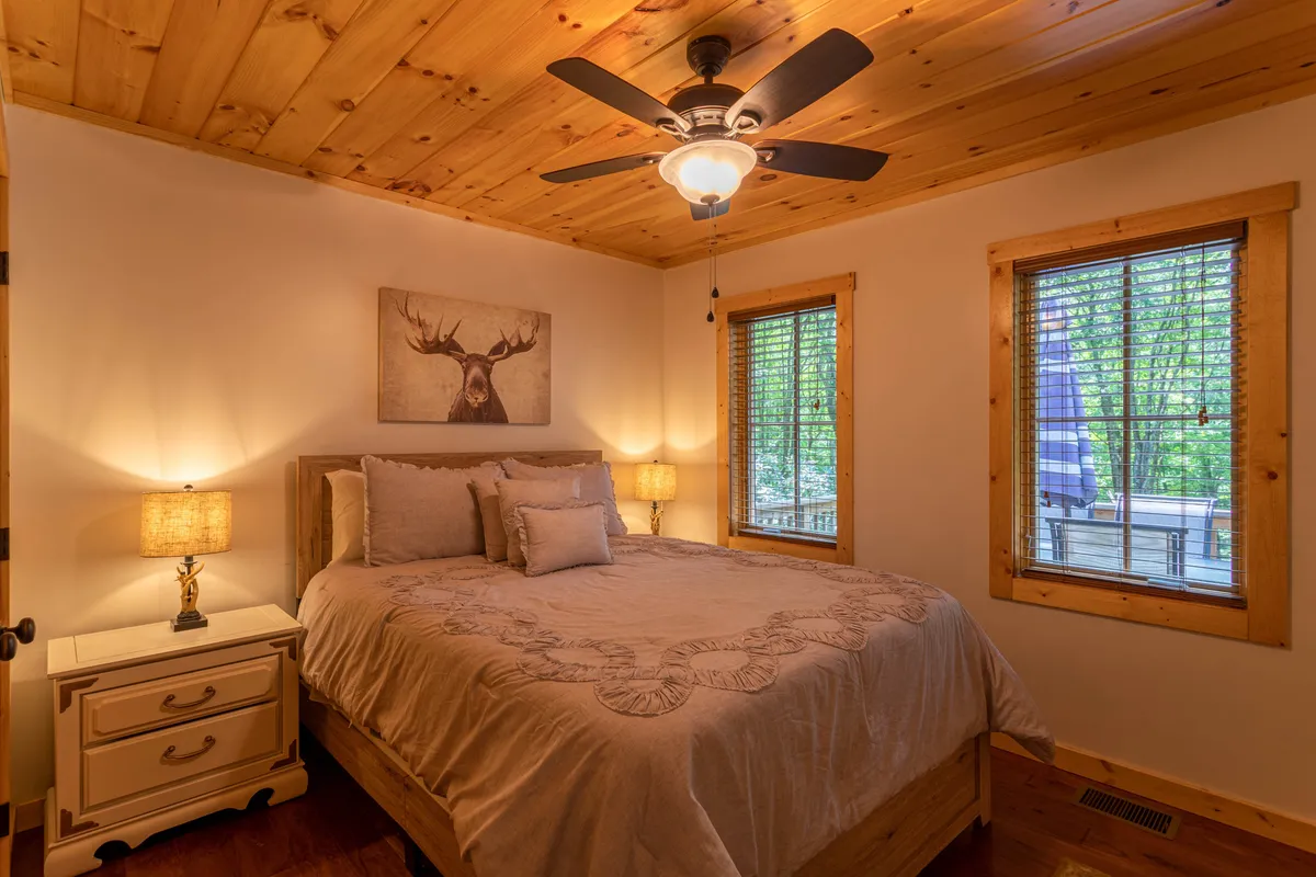 A cozy bedroom with a decorative wooden bed, moose artwork above the headboard, nightstands with lamps, and a window overlooking the outdoors. The room exudes warmth and invites guests to relax and unwind.