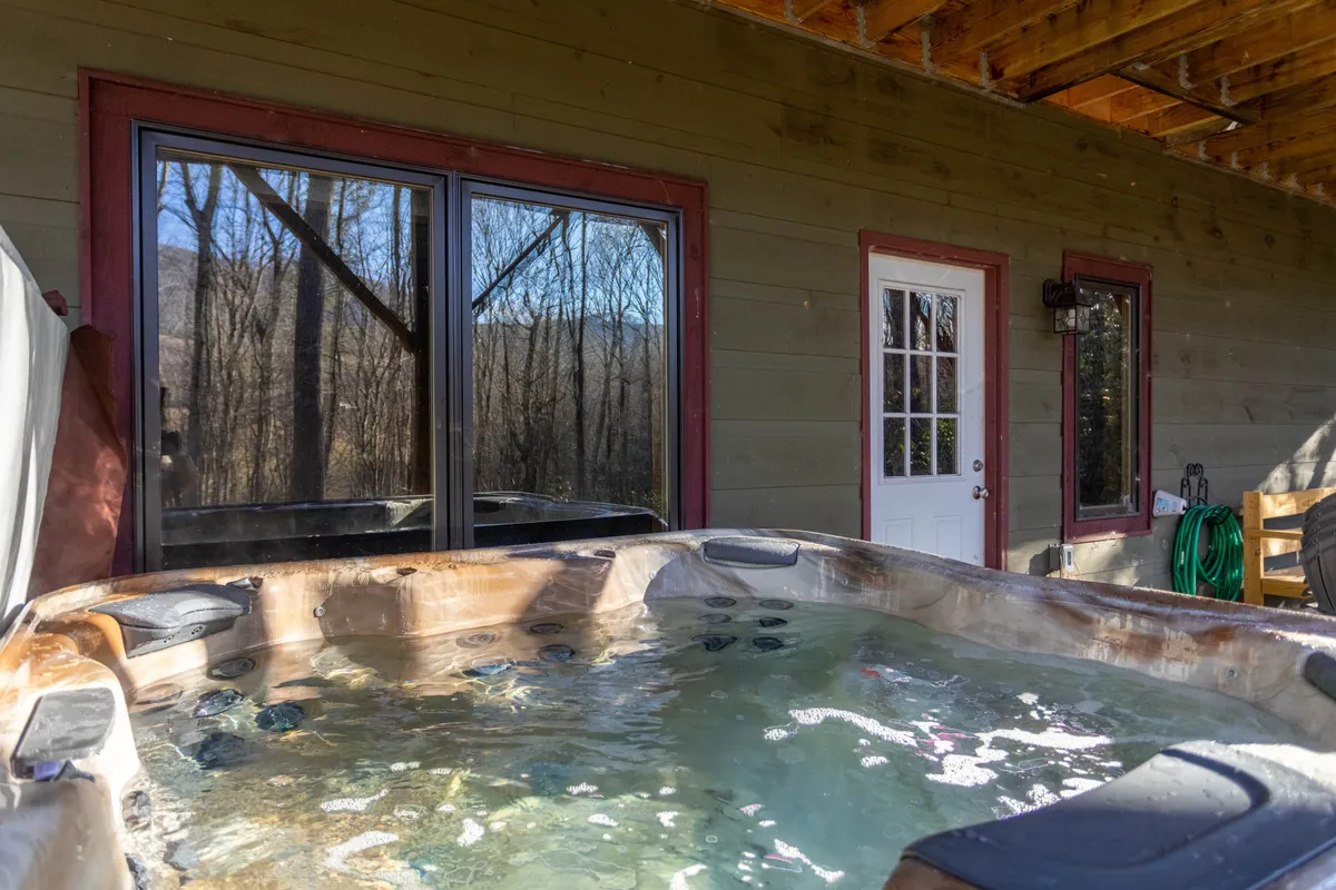 A serene outdoor hot tub tucked beside a wooden structure, with large windows reflecting the surrounding forest. The setting offers a private and peaceful spot for soaking while enjoying nature views, hinting at both relaxation and luxury.