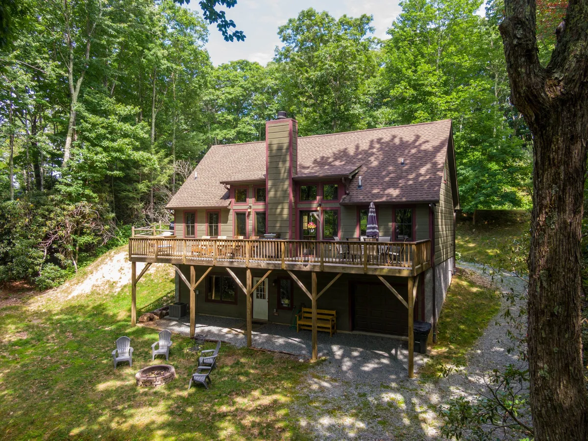 A cozy view of the home’s wooden deck featuring a fire pit, ample seating, and the backdrop of dense trees. The house’s exterior and deck suggest spaces for both relaxation and social gatherings, with an emphasis on enjoying the outdoors.