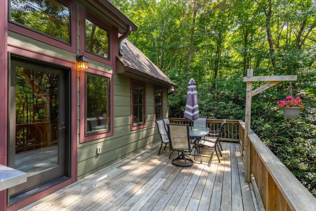 A spacious wooden deck with comfortable seating and umbrellas, surrounded by lush green foliage. The area is set up for outdoor dining, relaxation, and soaking in the natural mountain setting.