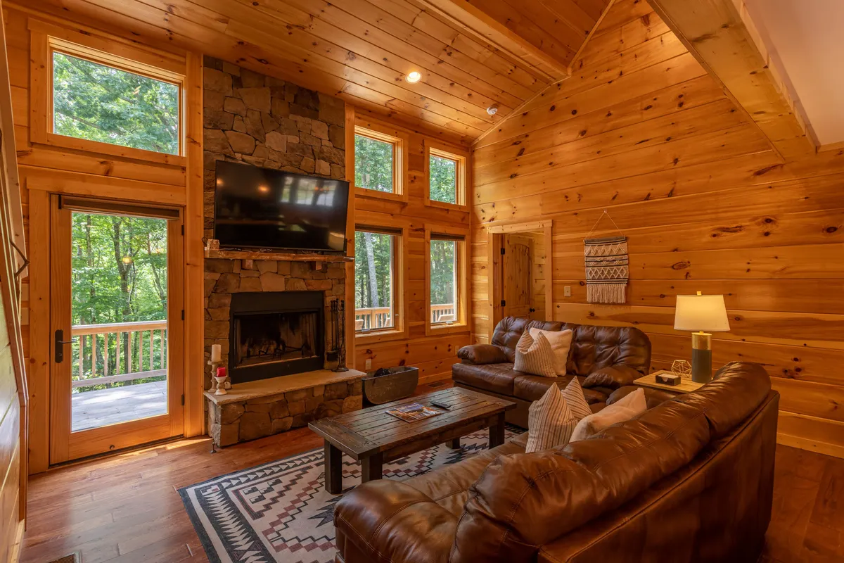 A welcoming great room with plush leather sofas arranged around a striking stone fireplace. Towering windows flood the space with natural sunlight and reveal a backdrop of lush woodland views. The setup is perfect for relaxing, socializing, or watching TV, with mountain-inspired décor adding warmth and charm.