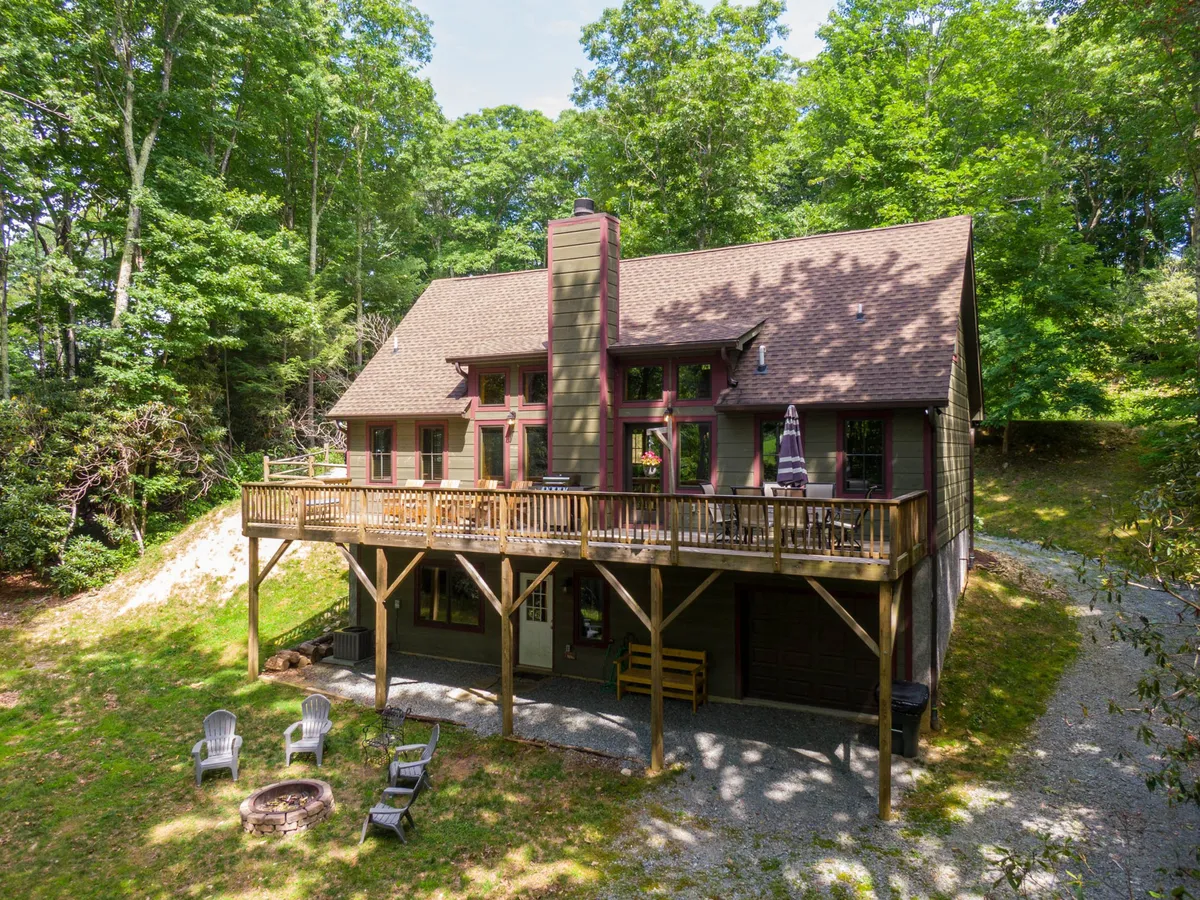 The hero image shows the exterior of a charming mountain lodge surrounded by lush greenery. A spacious wooden deck extends from the house, furnished for outdoor relaxation and offering sweeping sunset views. The deck overlooks a cozy fire pit area, perfect for gathering with friends and family, while the home's wall of large windows hints at bright, inviting interiors. The peaceful setting is nestled amidst the trees, creating a private, tranquil oasis.