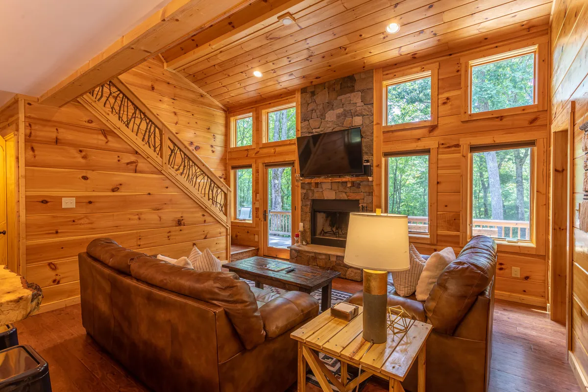 A spacious living area featuring a prominent fireplace, expansive windows offering sweeping views of the forest, and comfortable leather sofas with a wooden coffee table. A handsome wooden staircase hints at the open-concept flow between spaces.